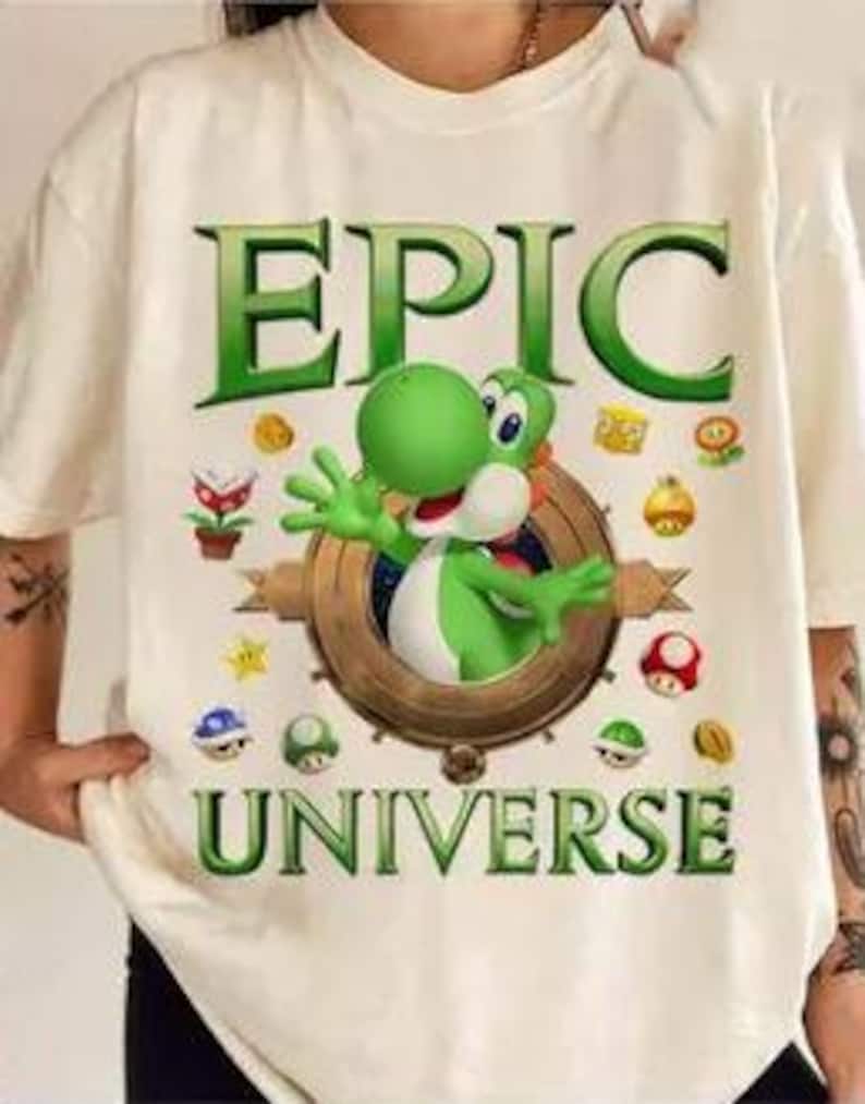 May include: White t-shirt with a green Yoshi character from the Super Mario video game series. The shirt has the text "EPIC UNIVERSE" printed on it.