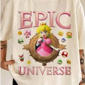 May include: White t-shirt with a pink and gold graphic of Princess Peach from the Super Mario Bros. video game series. The graphic includes the text "EPIC UNIVERSE" and features Princess Peach surrounded by various power-ups and coins.