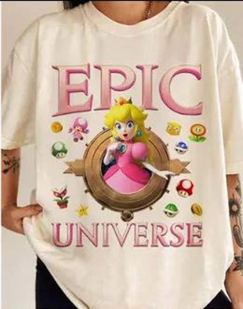 May include: White t-shirt with a pink and gold graphic of Princess Peach from the Super Mario Bros. video game series. The graphic includes the text "EPIC UNIVERSE" and features Princess Peach surrounded by various power-ups and coins.