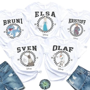 May include: Five white short-sleeved t-shirts featuring Disney character graphics. Each shirt displays a name, a checkered circle, the years 20 and 13, and a character: Bruni, Elsa, Kristoff, Sven, or Olaf. The Disney logo is printed below each circle.
