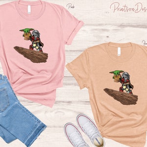 May include: Two t-shirts with a graphic print of a Mandalorian character holding a small green creature. The t-shirts are available in pink and heather peach.