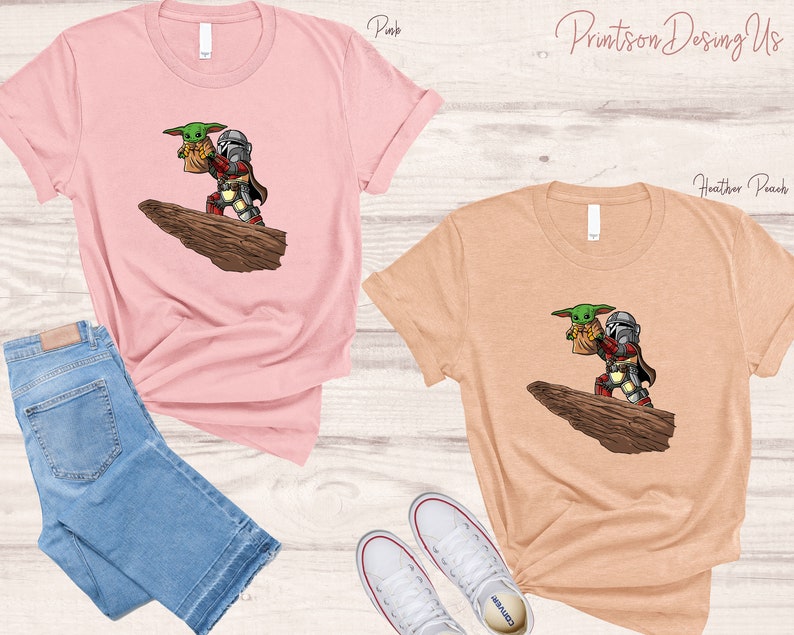 May include: Two t-shirts with a graphic print of a Mandalorian character holding a small green creature. The t-shirts are available in pink and heather peach.