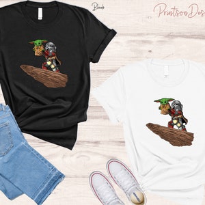 May include: Two t-shirts, one black and one white, featuring a cartoon image of a Mandalorian and a Grogu character from the Star Wars franchise. The Mandalorian is wearing his signature armor and holding Grogu in his arms. The image is set against a backdrop of a rocky cliff.