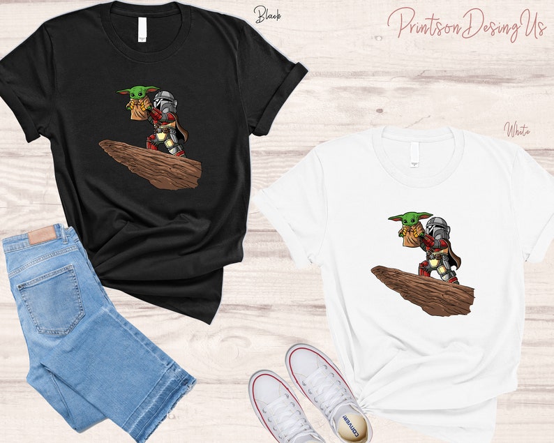May include: Two t-shirts, one black and one white, featuring a cartoon image of a Mandalorian and a Grogu character from the Star Wars franchise. The Mandalorian is wearing his signature armor and holding Grogu in his arms. The image is set against a backdrop of a rocky cliff.