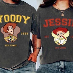 Puede incluir: Dos camisetas gris carb&oacute;n con personajes de Toy Story. Una camiseta muestra "WOODY" en amarillo, con la imagen de Woody, "EST 1995" y "TOY STORY". La otra camiseta muestra "JESSIE" en rojo, con la imagen de Jessie, "EST 1995" y "TOY STORY".
