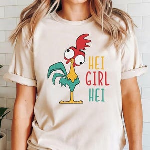 Pode incluir: Camiseta bege com um galo de desenho animado. O galo tem uma crista vermelha, asas turquesas e bico e pernas amarelas. O texto "HEI GIRL HEI" &eacute; impresso em amarelo, vermelho e turquesa. A camiseta tem mangas curtas.