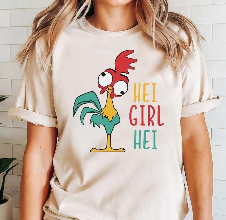 Pode incluir: Camiseta bege com um galo de desenho animado. O galo tem uma crista vermelha, asas turquesas e bico e pernas amarelas. O texto "HEI GIRL HEI" &eacute; impresso em amarelo, vermelho e turquesa. A camiseta tem mangas curtas.