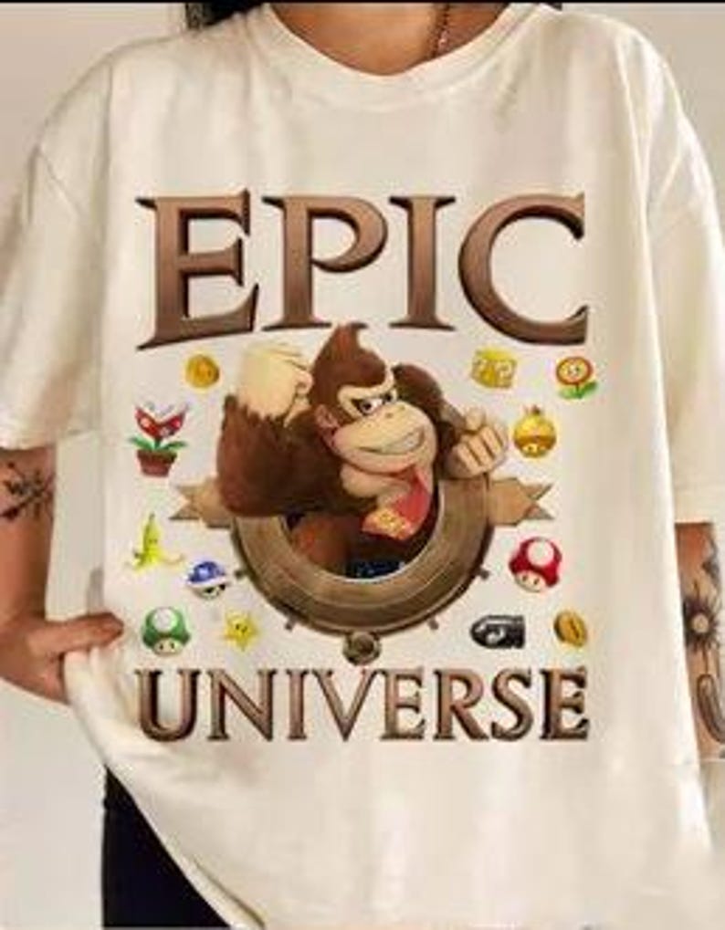 May include: White t-shirt with a brown cartoon gorilla wearing a red bandana and holding a banana. The gorilla is surrounded by video game icons and the words "EPIC UNIVERSE" are printed above and below the gorilla.