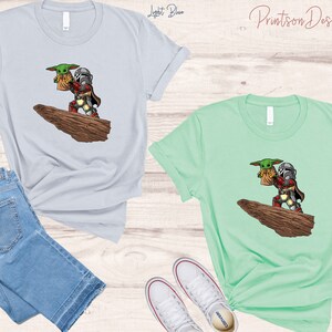 May include: Two t-shirts with a graphic print of a cartoon Mandalorian holding a baby Yoda. The t-shirt on the left is light blue and the t-shirt on the right is mint green.