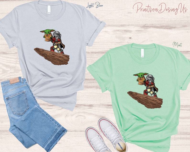 May include: Two t-shirts with a graphic print of a cartoon Mandalorian holding a baby Yoda. The t-shirt on the left is light blue and the t-shirt on the right is mint green.