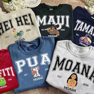 May include: A collection of crewneck sweatshirts in various colors, each featuring a different design and the word "Moana" or a character's name in bold, arched lettering. The sweatshirts have cartoon illustrations and the year "2016".