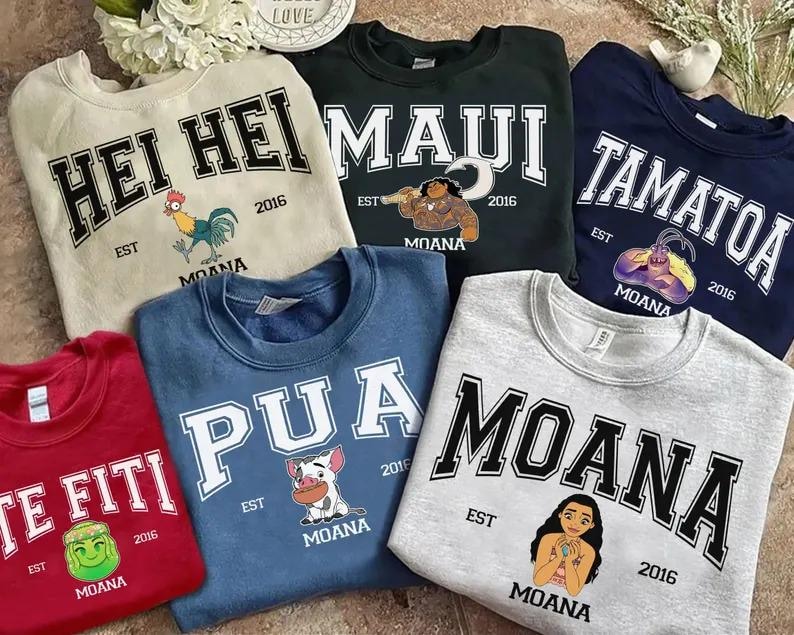 May include: A collection of crewneck sweatshirts in various colors, each featuring a different design and the word "Moana" or a character's name in bold, arched lettering. The sweatshirts have cartoon illustrations and the year "2016".