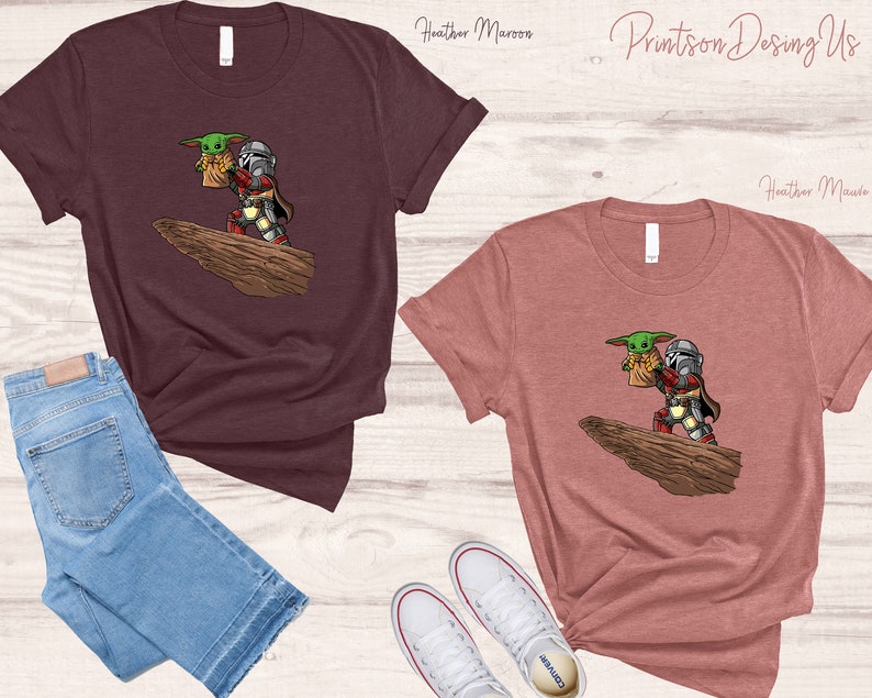 May include: Two t-shirts with a graphic print of a green alien character being carried by a Mandalorian character. The t-shirts are in heather maroon and heather mauve colors.