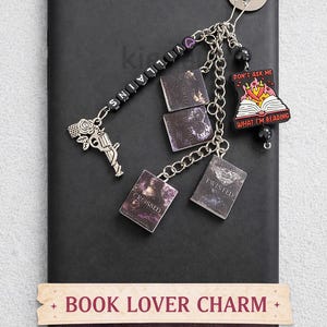 Op de afbeelding: Een zwarte Kindle met een zilveren kettingbedel. De bedel bevat miniatuur boekenhangers, een pistool- en rozenbedel en een bedel met de tekst "DON'T ASK ME WHAT I'M READING". De tekst "VILLAINS" is gespeld op zwarte kralen. De tekst "BOOK LOVER CHARM" en "KINDLE CHAIN" zijn ook zichtbaar.