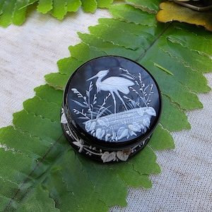 Puede incluir: Una caja de baratijas redonda negra con un diseño de garza blanca. La garza se encuentra en una zona pantanosa con plantas y agua. La caja tiene un diseño floral decorativo alrededor de los lados. La caja está sobre una hoja verde.