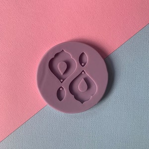 May include: A lavender-coloured, circular silicone mould featuring two ornate teardrop-shaped designs. Each design has a central teardrop shape and smaller, circular accents. The mould is set against a backdrop of pink and light blue paper.