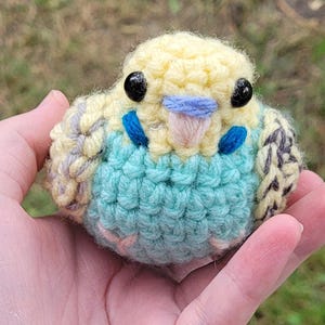 May include: A handmade crochet bird in yellow, blue, and gray. The bird has black bead eyes, a blue beak, and blue accents. The body is primarily blue, with yellow and gray detailing. A unique, handcrafted item.