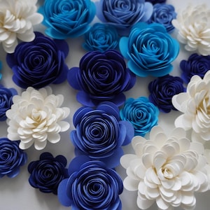 May include: An assortment of paper flowers in shades of white, blue, and navy. The arrangement includes various sizes of roses and other layered floral designs, creating a textured and colorful display.