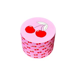 May include: Pink metal grinder with a cherry design. The top of the grinder has a single cherry with a stem and leaves. The sides of the grinder are covered in a repeating pattern of cherries.