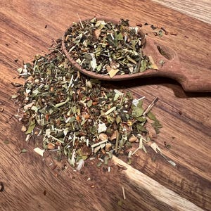 May include: A wooden spoon filled with a mix of dried herbs and spices, including green leaves, brown stems, and orange pieces. The spoon rests on a wooden cutting board with a natural wood grain pattern. The tea blend is scattered around the spoon.