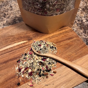 May include: A wooden spoon scoops a pile of dried herbs and flowers, including rose petals, berries, and dried leaves. The herbs are in a brown paper bag with a clear window.
