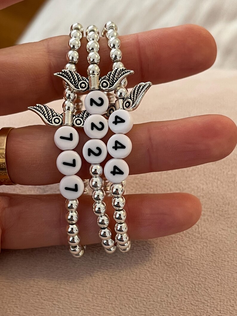 Pu&ograve; includere: Tre braccialetti di perline con perline e ciondoli color argento. Ogni braccialetto ha perline di lettere bianche che compongono i numeri: 777, 222 e 444. I ciondoli sono ali d'angelo.