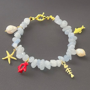 May include: A beaded bracelet featuring light blue stones and gold-colored charms. Charms include a starfish, coral, fish, and fishbone. The bracelet also has two white pearl accents and a gold clasp.