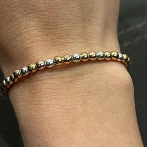 May include: A bracelet with a mix of gold and silver-coloured beads. The beads are small and round, strung together to create a flexible accessory. The bracelet is worn on a wrist. The beads reflect light, creating a subtle shimmer.