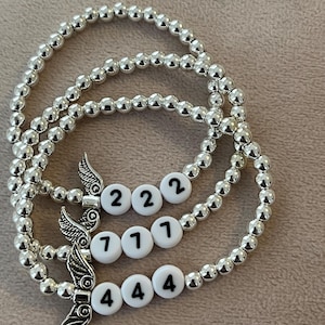 Pu&ograve; includere: Tre braccialetti con perline argentate. Ogni braccialetto presenta perline bianche con numeri neri e un ciondolo a forma di ala argentata. I numeri sui braccialetti sono 222, 711 e 444.