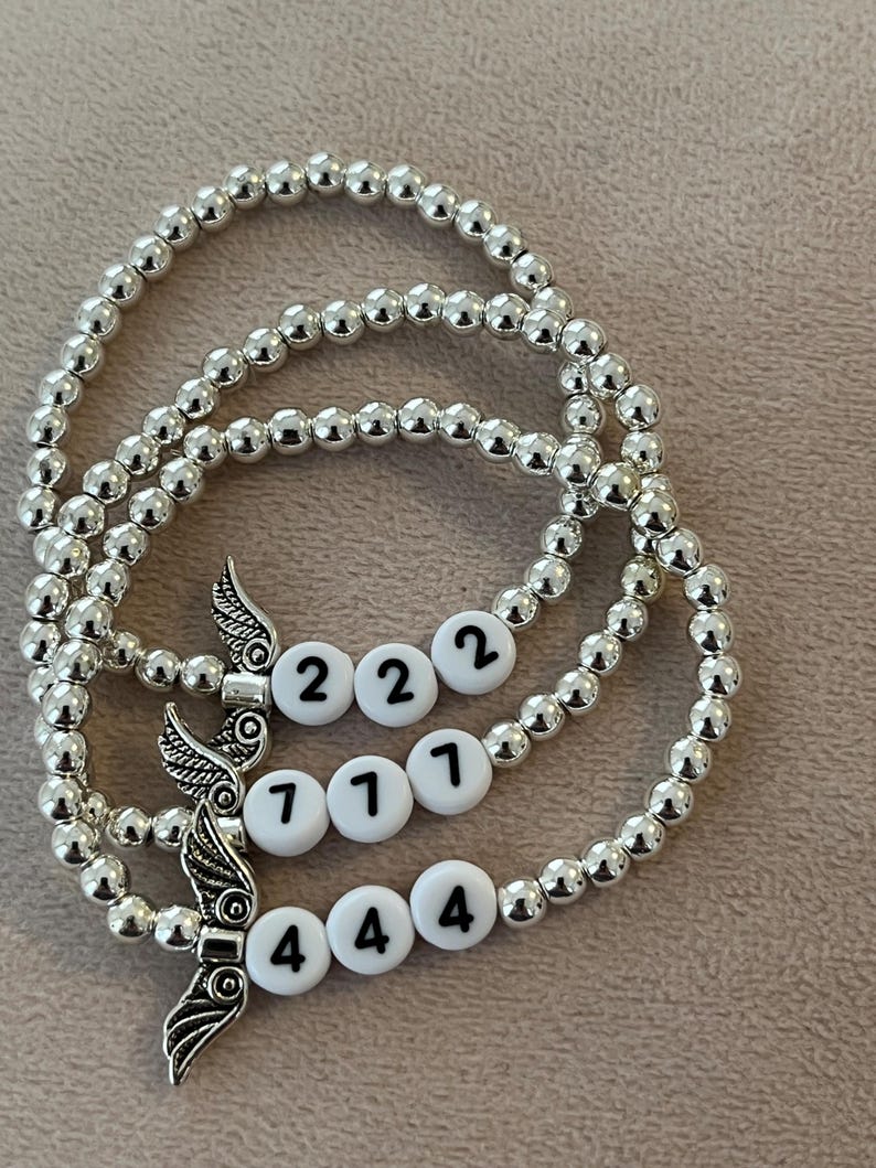 Pu&ograve; includere: Tre braccialetti con perline argentate. Ogni braccialetto presenta perline bianche con numeri neri e un ciondolo a forma di ala argentata. I numeri sui braccialetti sono 222, 711 e 444.