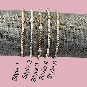 Peut inclure: Cinq styles différents de bracelets en perles, présentés sur une surface en tissu gris. Les bracelets sont faits de petites perles dorées et argentées. Les bracelets sont étiquetés "Style 1" à "Style 5".