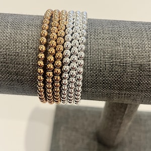 May include: A set of six stretch bracelets featuring small, round, metallic beads. Three of the bracelets are finished in a warm gold tone, whilst the other three are finished in a bright silver tone, all arranged in a row on a display stand.