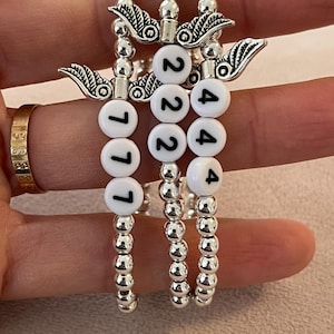 Pu&ograve; includere: Tre braccialetti con perline argentate. Ogni braccialetto presenta perline bianche con numeri neri e un ciondolo argentato con dettagli di ali. I numeri sui braccialetti sono 777, 222 e 444.