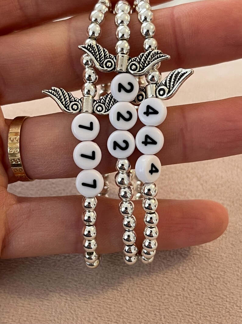 Pu&ograve; includere: Tre braccialetti con perline argentate. Ogni braccialetto presenta perline bianche con numeri neri e un ciondolo argentato con dettagli di ali. I numeri sui braccialetti sono 777, 222 e 444.