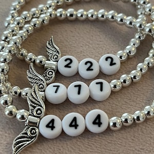 Pu&ograve; includere: Braccialetti con perline d'argento con perline bianche che mostrano i numeri 222, 777 e 444. Ogni braccialetto presenta un ciondolo a forma di ala d'argento. I braccialetti sono esposti su una superficie rosa chiaro.