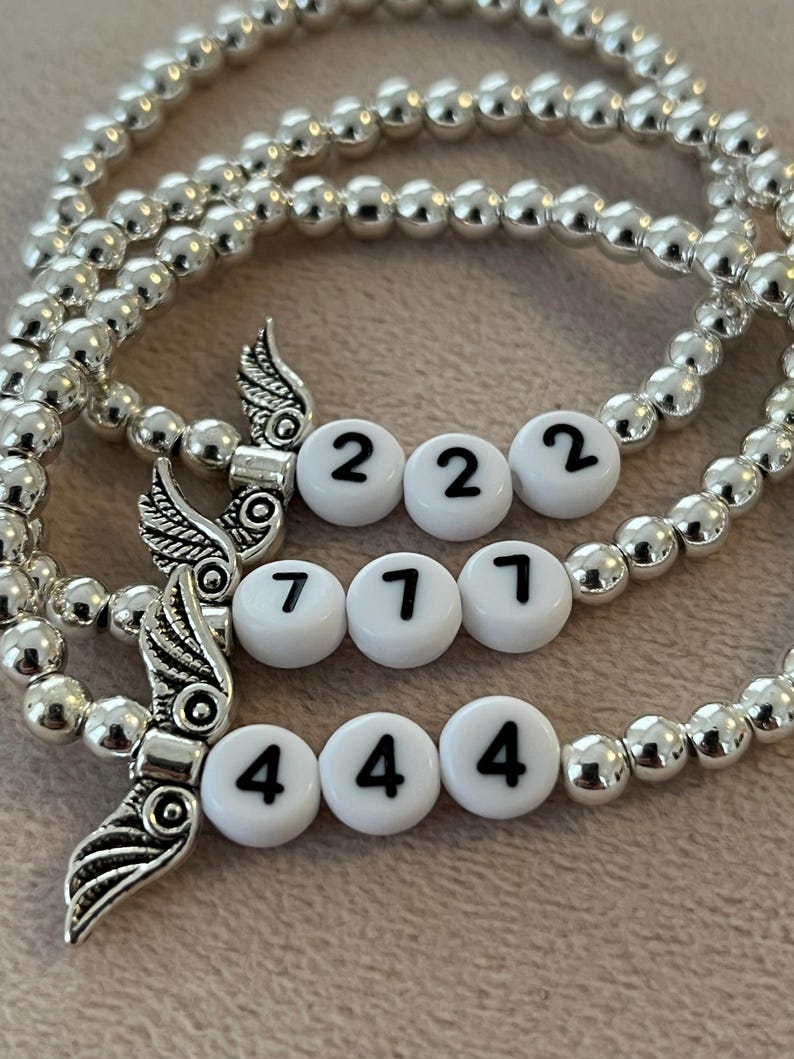 Pu&ograve; includere: Braccialetti con perline d'argento con perline bianche che mostrano i numeri 222, 777 e 444. Ogni braccialetto presenta un ciondolo a forma di ala d'argento. I braccialetti sono esposti su una superficie rosa chiaro.