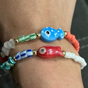 May include: Two beaded bracelets with fish-shaped charms. One bracelet features a blue fish with white polka dots, green and gold beads, and coral beads. The other bracelet has a red fish charm, blue and white beads, and turquoise stones.