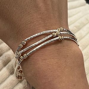 May include: A silver bracelet with three strands, each adorned with small gold beads. The bracelet has a textured, hammered appearance on the silver strands. The gold beads are evenly spaced along the bracelet's circumference, creating a delicate and elegant design.