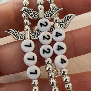 Pu&ograve; includere: Tre braccialetti di perline con perline e ciondoli argentati. Ogni braccialetto ha perline rotonde bianche con numeri neri: 111, 222 e 444. I braccialetti sono tenuti insieme da ciondoli argentati con un design ad ala.