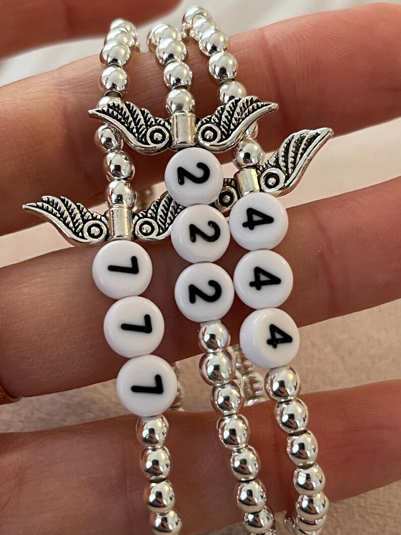 Pu&ograve; includere: Tre braccialetti di perline con perline e ciondoli argentati. Ogni braccialetto ha perline rotonde bianche con numeri neri: 111, 222 e 444. I braccialetti sono tenuti insieme da ciondoli argentati con un design ad ala.