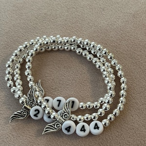 Pu&ograve; includere: Tre braccialetti di perline d'argento con perline di lettere bianche che compongono numeri. Ogni braccialetto ha un ciondolo a forma di ala d'argento. I braccialetti sono esposti su una superficie di colore neutro.