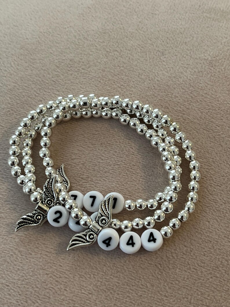 Pu&ograve; includere: Tre braccialetti di perline d'argento con perline di lettere bianche che compongono numeri. Ogni braccialetto ha un ciondolo a forma di ala d'argento. I braccialetti sono esposti su una superficie di colore neutro.