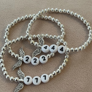 Pu&ograve; includere: Tre braccialetti con perline argentate. Ogni braccialetto presenta un ciondolo con dettagli alati e perline bianche con i numeri 222, 444 e 777. I braccialetti sono esposti su una superficie dai toni neutri.