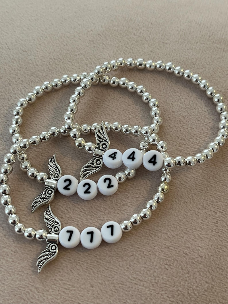 Pu&ograve; includere: Tre braccialetti con perline argentate. Ogni braccialetto presenta un ciondolo con dettagli alati e perline bianche con i numeri 222, 444 e 777. I braccialetti sono esposti su una superficie dai toni neutri.