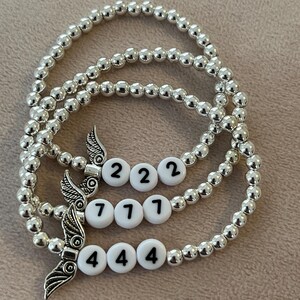 Pu&ograve; includere: Tre braccialetti con perline argentate. Ogni braccialetto presenta perline bianche con i numeri 222, 771 e 444. Ogni braccialetto ha anche un ciondolo a forma di ala argentata. I braccialetti sono esposti su una superficie di colore neutro.