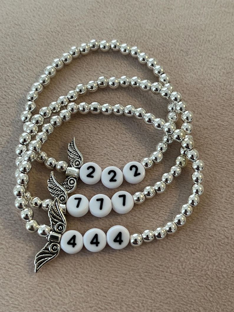 Pu&ograve; includere: Tre braccialetti con perline argentate. Ogni braccialetto presenta perline bianche con i numeri 222, 771 e 444. Ogni braccialetto ha anche un ciondolo a forma di ala argentata. I braccialetti sono esposti su una superficie di colore neutro.