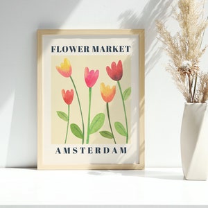 May include: A framed art print with a watercolor illustration of tulips in shades of red, pink, and yellow. The print reads "FLOWER MARKET AMSTERDAM" in navy blue text. A white vase with dried pampas grass sits to the right.