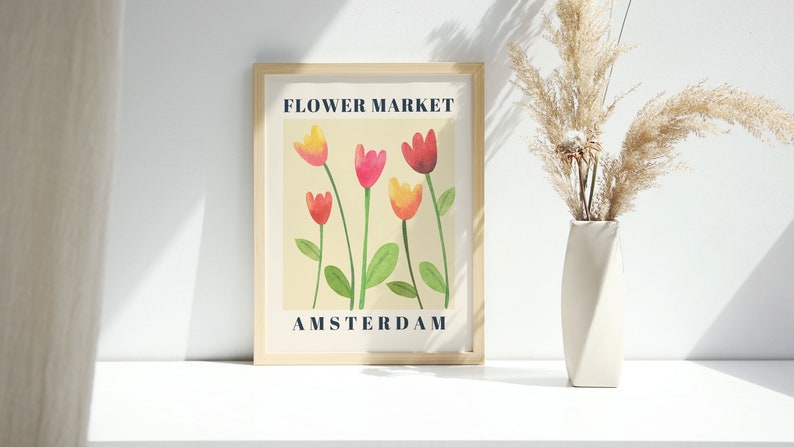 May include: A framed art print with a watercolor illustration of tulips in shades of red, pink, and yellow. The print reads "FLOWER MARKET AMSTERDAM" in navy blue text. A white vase with dried pampas grass sits to the right.
