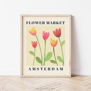 May include: A framed art print featuring a watercolor illustration of tulips in shades of red, pink, and yellow. The text "FLOWER MARKET AMSTERDAM" is displayed in a navy blue font against a cream background.