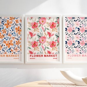 May include: Three framed floral prints with the text "FLOWER MARKET" and the city and country of origin below. The prints feature different floral patterns in shades of pink, orange, and blue.